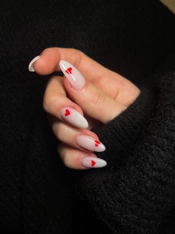 Close-up of a woman's hand with acrylic nails painted white with red heart designs, showcasing modern nail art.